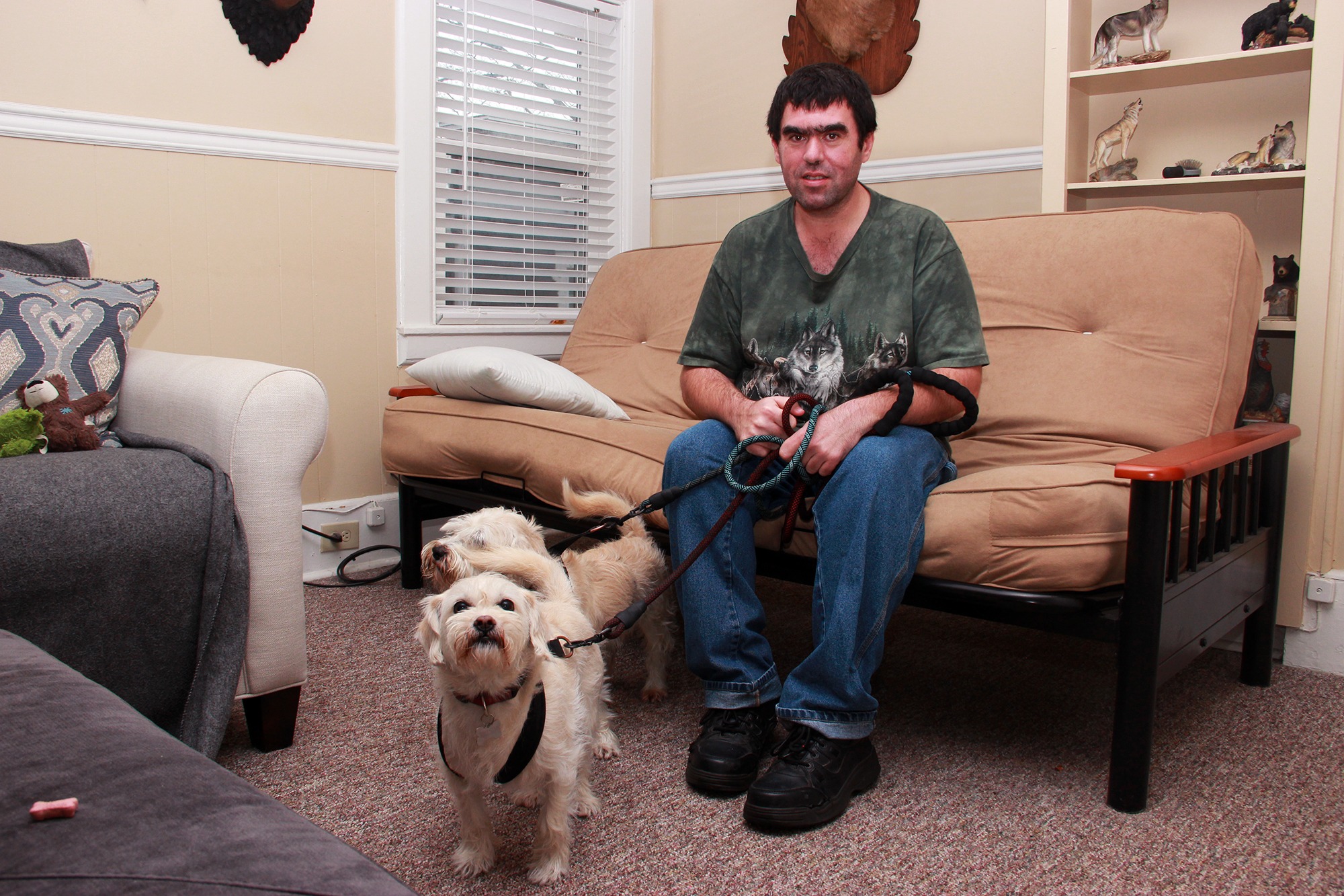 Patrick Toomey sitting on a futon couch in his living room with his two small terrier dogs on a leash.